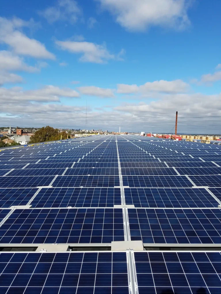 Aerial view of a large commercial solar panel installation under a blue sky, showcasing numerous solar panels aligned on a rooftop, emphasizing clean energy solutions by BD Electrical Services.