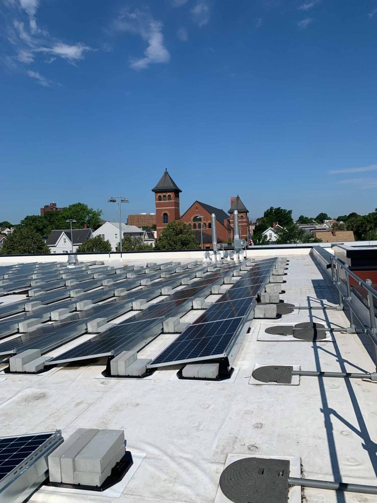 Solar panel installation on the roof of East Somerville Community School in Somerville, MA, showcasing multiple solar panels under a clear blue sky, with historic buildings visible in the background.