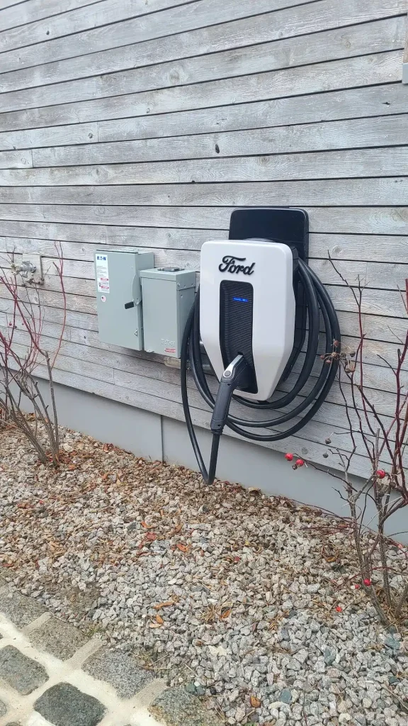 Ford electric vehicle charger mounted on a wall with vehicle-to-home backup integration, surrounded by gravel landscaping and shrubbery, showcasing clean energy solutions for Massachusetts businesses.