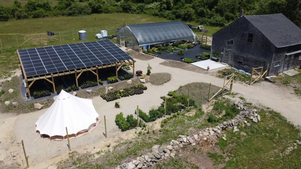 Aerial view of a small business solar installation featuring solar panels on a wooden structure, surrounded by a garden, greenhouse, and tent, showcasing sustainable energy solutions for commercial use.