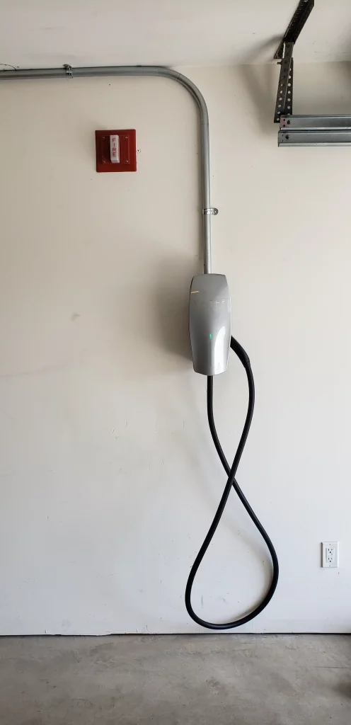 Tesla electric vehicle charging station mounted on a wall, with a coiled charging cable, in a garage setting, highlighting clean energy solutions for Massachusetts businesses.