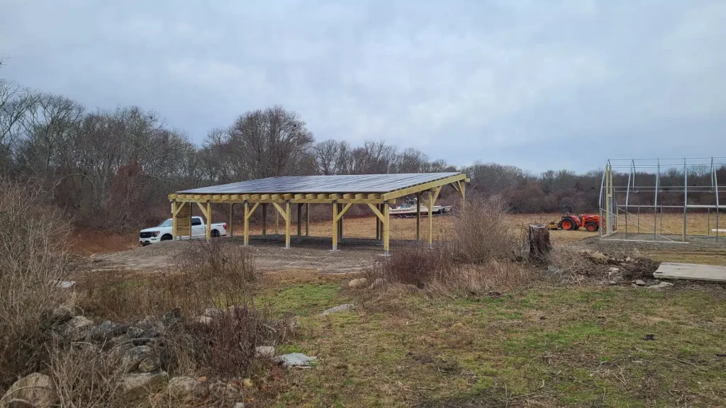 Residential solar pergola installation in Westport, MA, featuring solar panels on a wooden structure, surrounded by natural landscape and equipment.